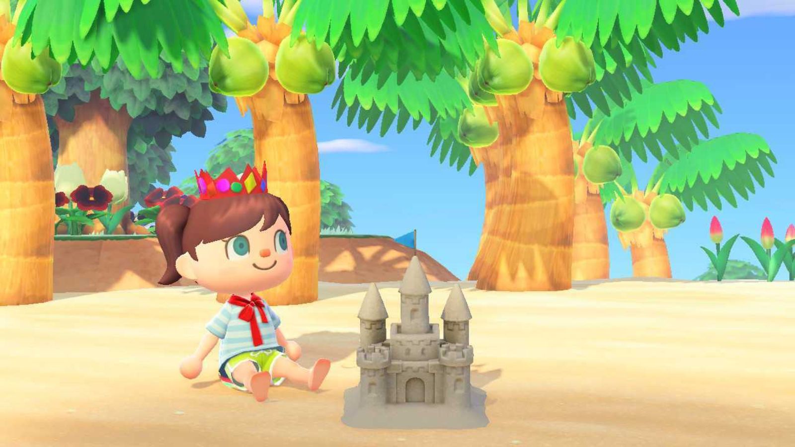 Animal Crossing New Horizons. The player is sat by a sandcastle on the beach while wearing a crown and a cape. There are coconut trees in the background. The player is on the left.