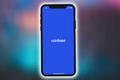 Image of the Coinbase Logo on a blue background on a phone, against a blurred blue/orange background.