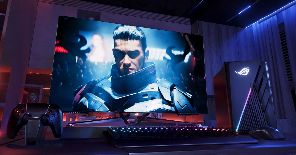 A monitor in a dark purple and blue-lit room with a video game character in armour on the display next to a PC.