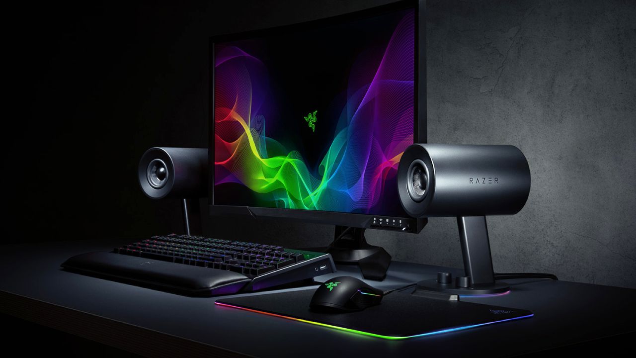 A black monitor with a multicolour pattern on the display next to a set of black speakers and an RGB keyboard.