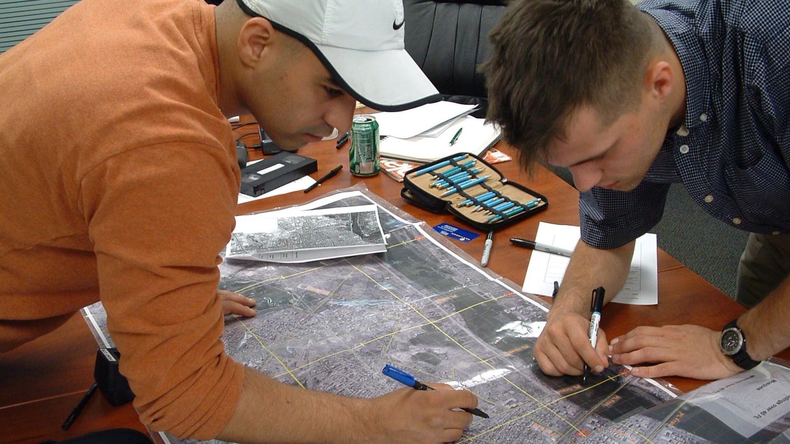 Two men stand over a map on a table. Pens in hand.
