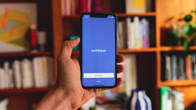 Image of hand holding a phone displaying the Coinbase application, against blurred background of books.