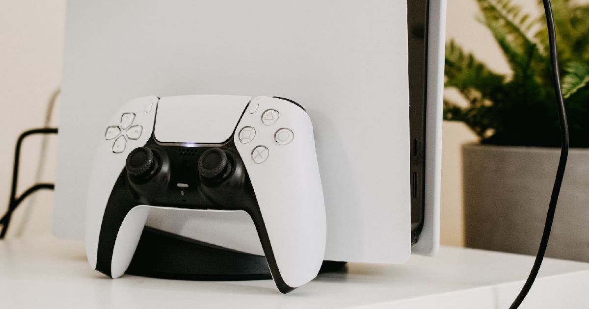 A white and black PS5 next to a house plant with a controller leaning against it.