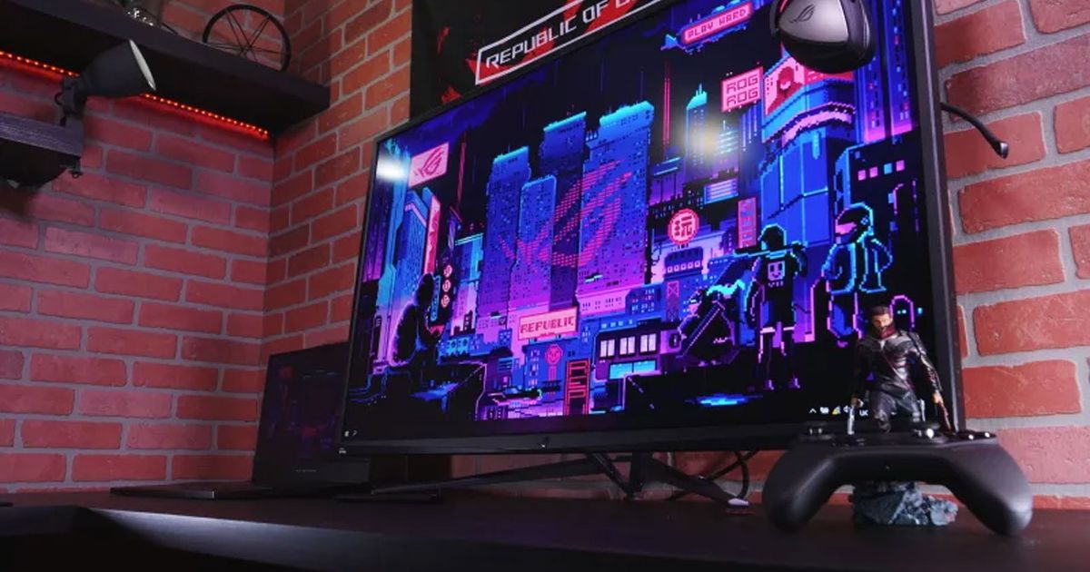 A black monitor with a purple and blue city scene on the display sat on a desk with a black Xbox controller in front of a red brick wall.