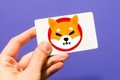 Shiba Inu logo on a card, held by a hand against a purple background, after a SHIB whale bough $250,000 worth of gift cards.