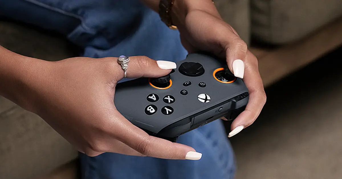 Someone with painted white finger nails using a grey SCUF Xbox controller featuring black buttons and orange trim around the joysticks.