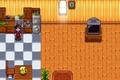 Stardew Valley. The player is standing on the left of the image in their kitchen. The kitchen has black and white tiles on the ground. The player is stood in front of their silver stove and to the left of a silver fridge. There is a wooden table and chair at the bottom of the image. 