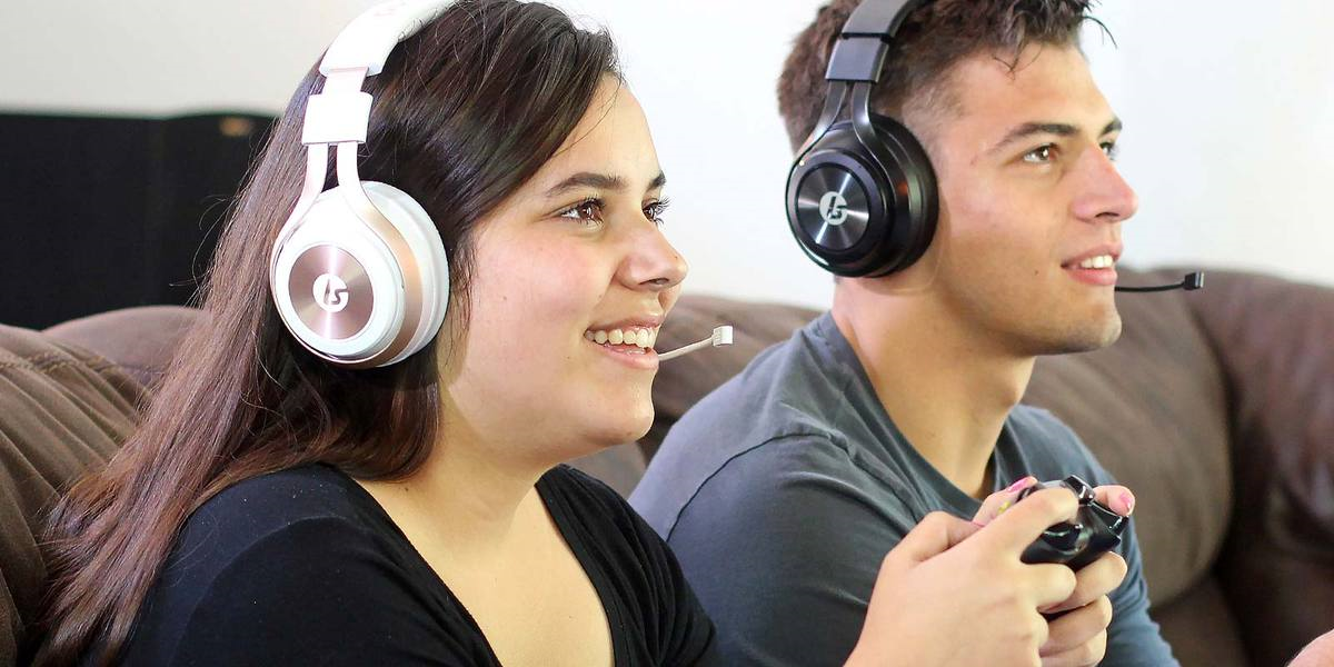 LucidSound LS35X Wireless Gaming Headset - Promotional image showing two adults playing games, woman is wearing the Rose Gold/White LS35X headset. Man is wearing a Black LS35X headset. 