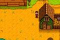 Stardew Valley. The player is stood to the left of their basic coop. The coop is on the right. It's green with a brown door and brown roof. There is a small brown fence holding hay and a bucket of water on the right of the coop.