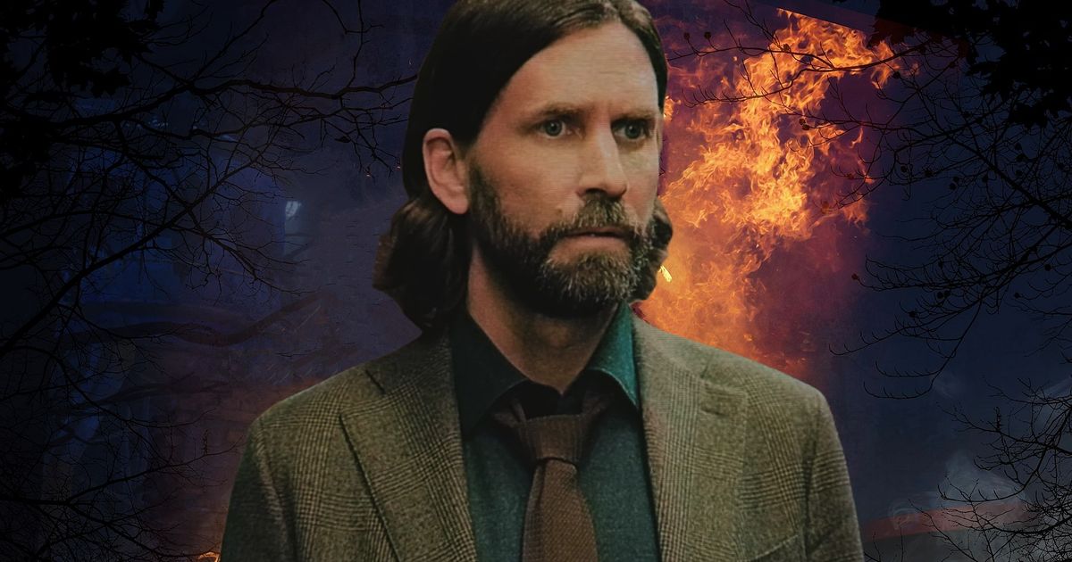 Man with long black hair and a beard in a green suit, stood in front of a fire