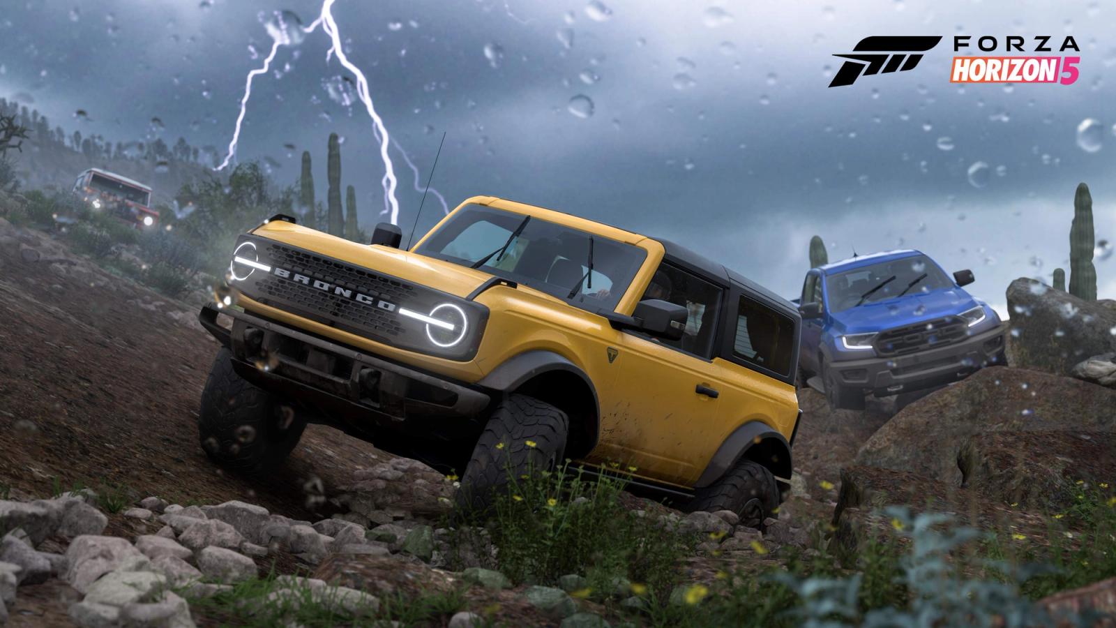 A yellow Ford Bronco drives across a dirt track towards the camera as a blue car is in pursuit. Lightning flashes in the background