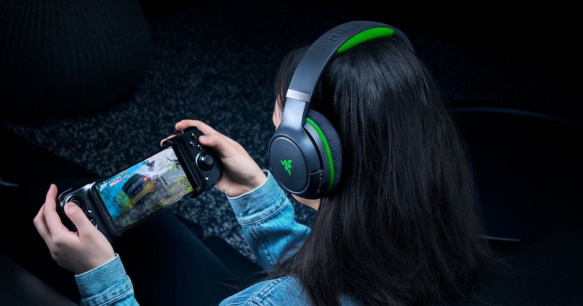 Someone with a black and green over-ear headset on playing a racing game on their phone using a controller.