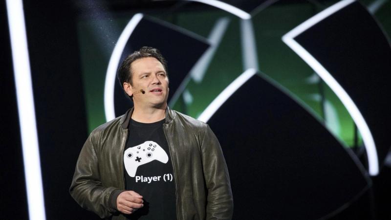 Phil spencer wearing xbox console controller black t shirt speaking on stage with xbox logo in background