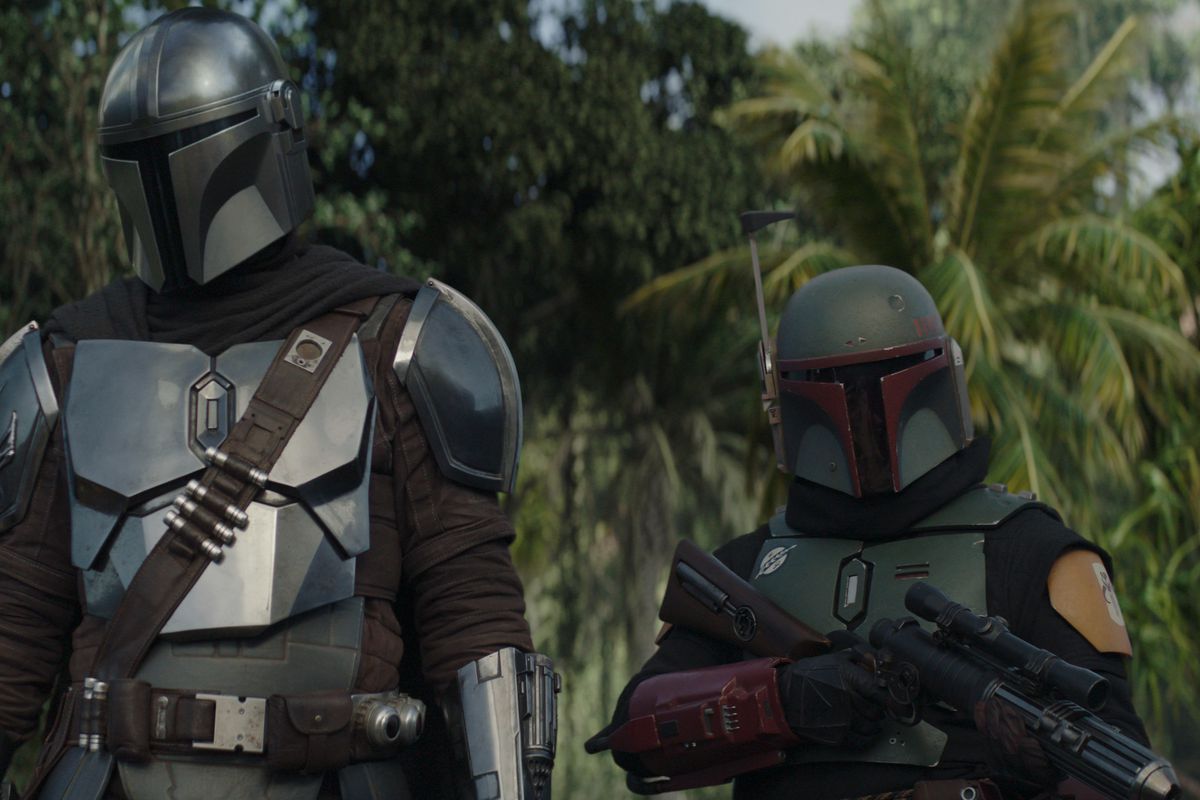 The Mandalorian and Boba Fett are standing next to each other in a forest.