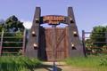 Jurassic World Evolution 2. The image shows the iconic Jurassic Park entrance gates. Jurassic Park is written across the top of the gate and the doors are wooden.
